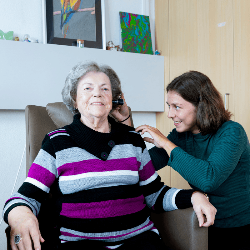 Een glimlachende, oudere vrouw met grijs haar en een gestreepte trui zit in een stoel en kijkt in de camera. Naast haar staat een jongere vrouw in een donkergroene trui, die in haar oor kijkt.