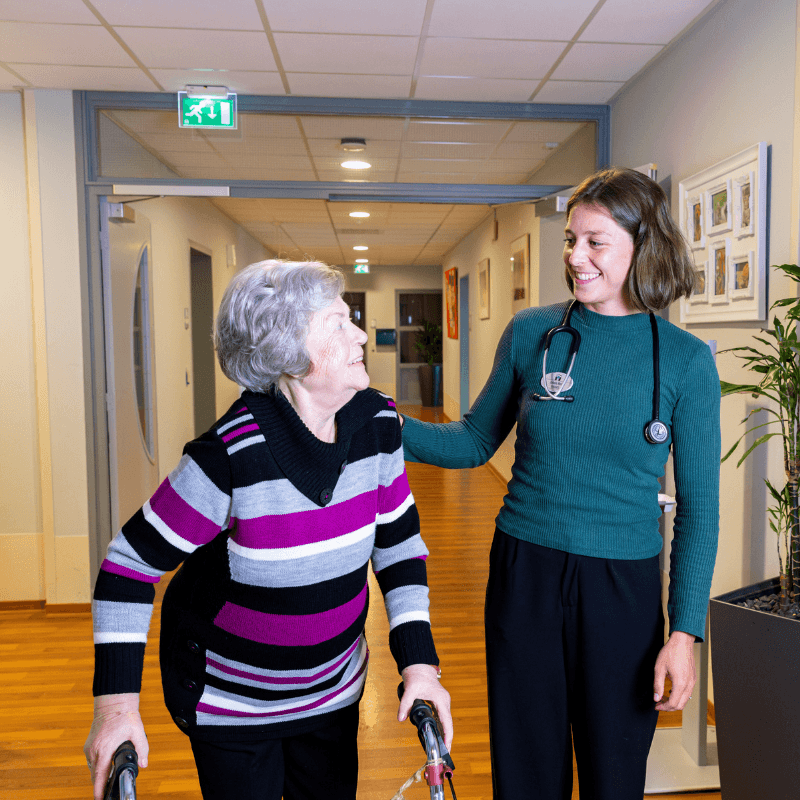 Een glimlachende jonge vrouw met een stethoscoop om haar nek helpt een oudere vrouw die een looprek gebruikt. De jonge vrouw heeft haar hand op de schouder van de oudere vrouw.