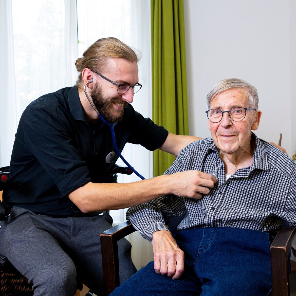 Een man met een knotje en een stethoscoop luistert naar het hart van een oudere, zittende man met een geruit bloesje.