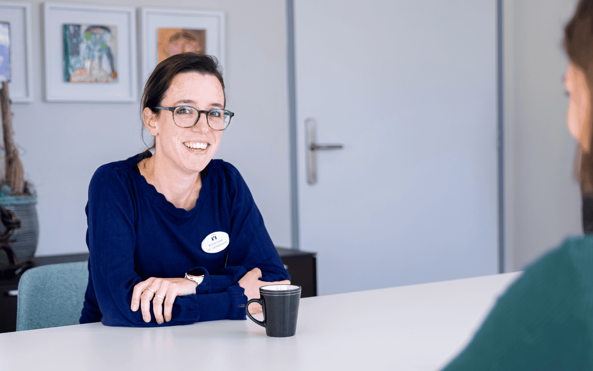 Lachende medewerker van MeanderGroep zit aan een tafel met een mok voor zich.