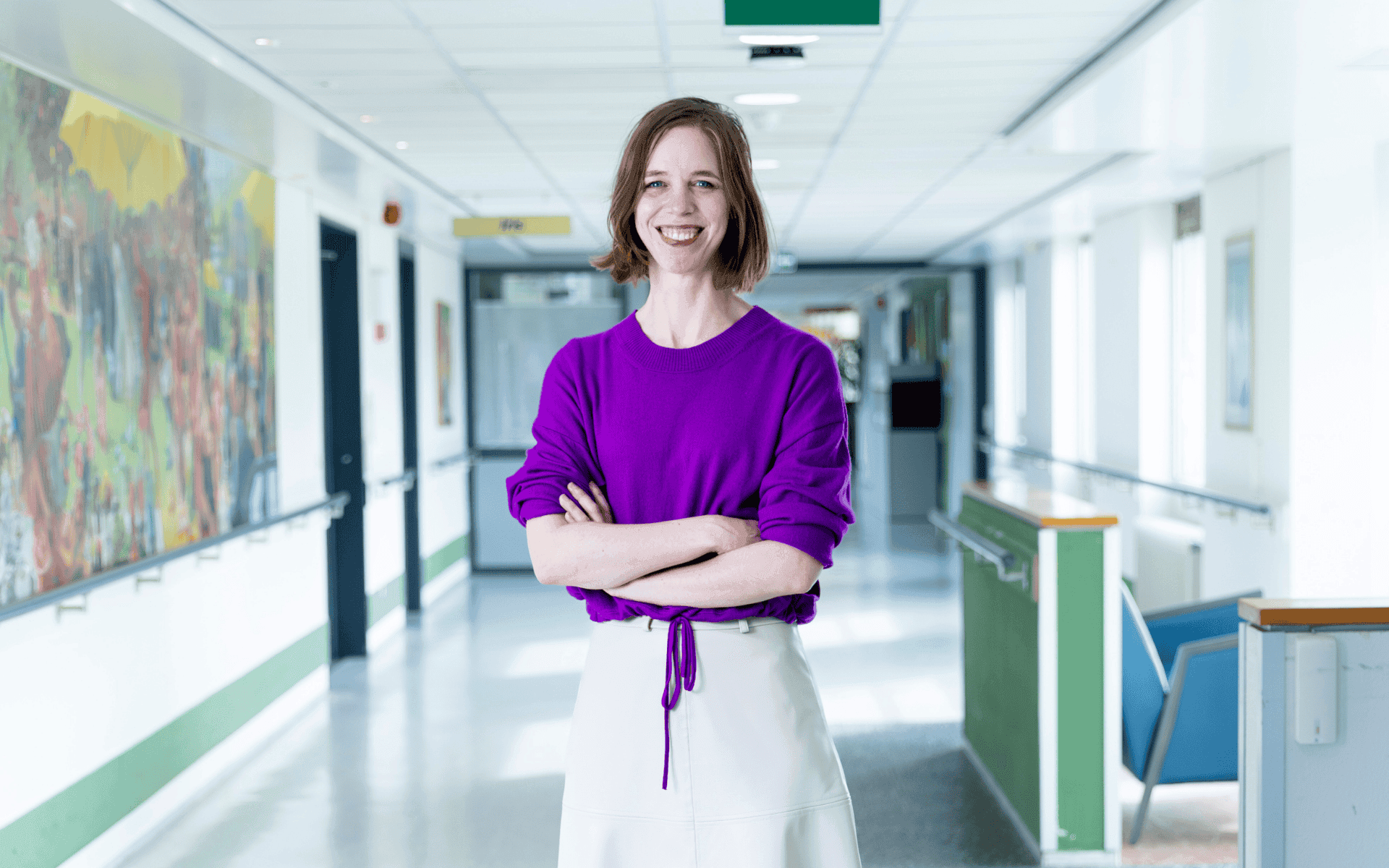 Een jonge vrouw met kort bruin haar staat lachend in een ziekenhuisgang. Ze draagt een felpaarse trui en een lichte rok en staat met haar armen gekruist.