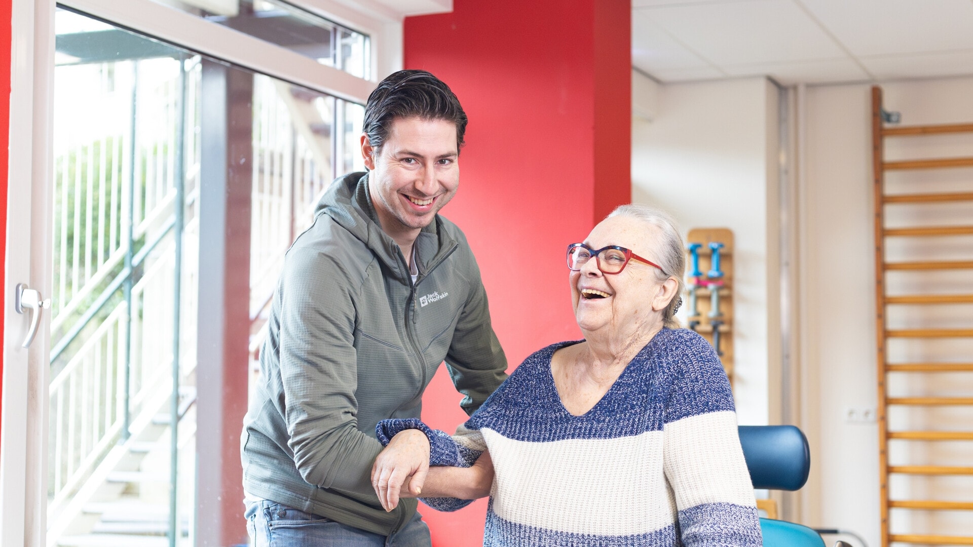 Een man met donker haar glimlacht en ondersteunt een lachende oudere vrouw die een gestreepte trui en een bril met rood montuur draagt. Ze bevinden zich in een fysiotherapieruimte.