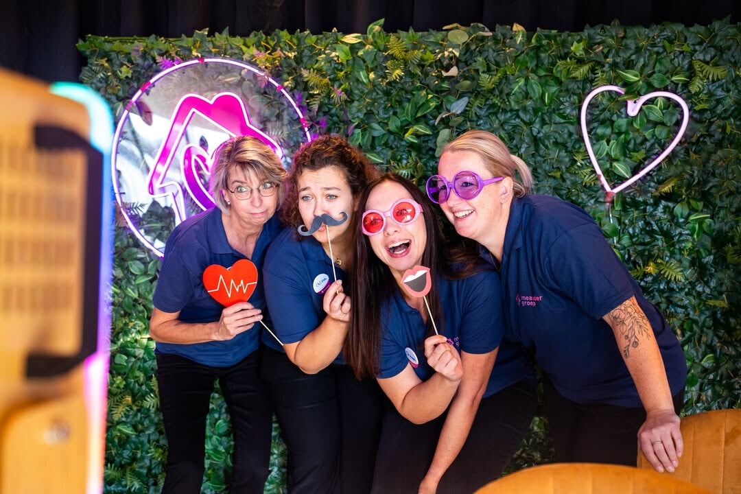 Vier vrouwelijke medewerkers van Meandergroep poseren voor een bladerwand op het personeelsfeest.
