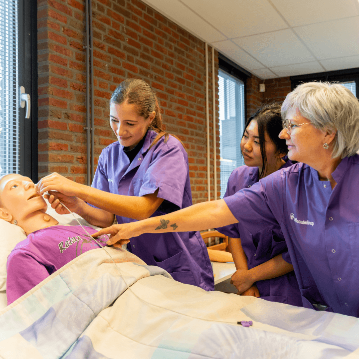 Oudere zorgmedewerker met twee jongere zorgmedewerkers staan naast een bed met een medische oefenpop.