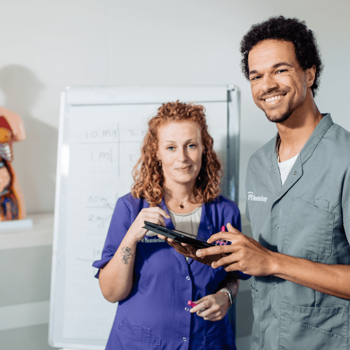 Twee medische professionals, een vrouw en een man, in gesprek met een tablet in een klaslokaalomgeving met een whiteboard en een anatomisch model op de achtergrond.