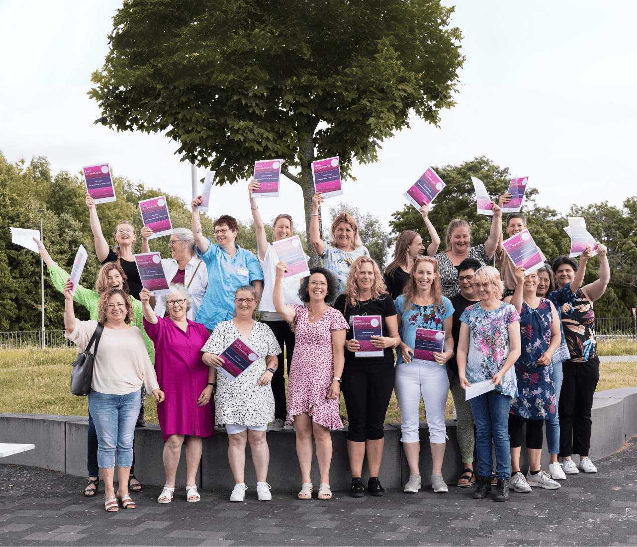 Een grote groep volwassen vrouwen staan buiten met hun armen in de lucht, elk met een paars en roze diploma.