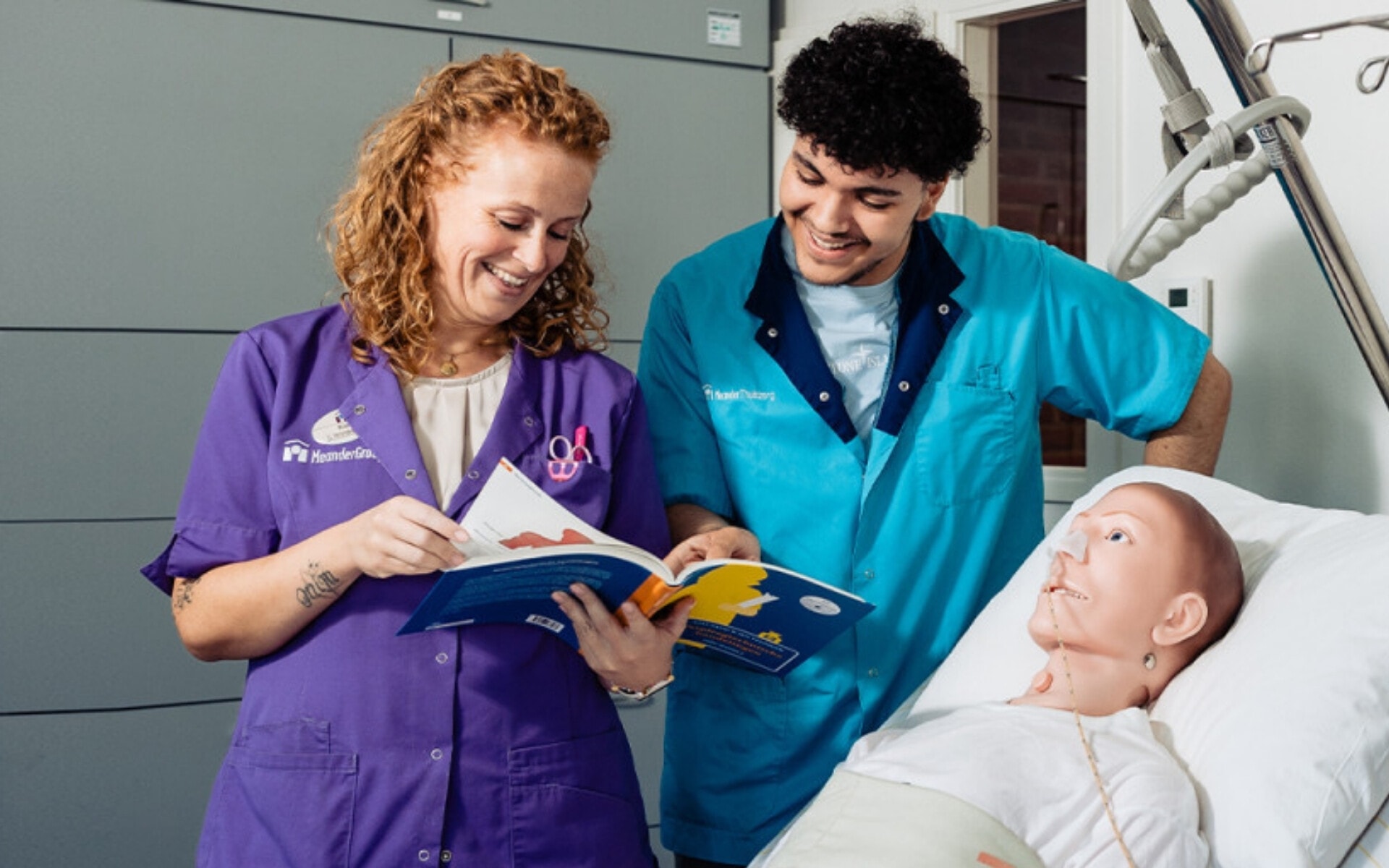 Twee vrolijke zorgmedewerkers, een man en een vrouw, staan naast een bed en bestuderen samen een boek. Op het bed ligt een pop.