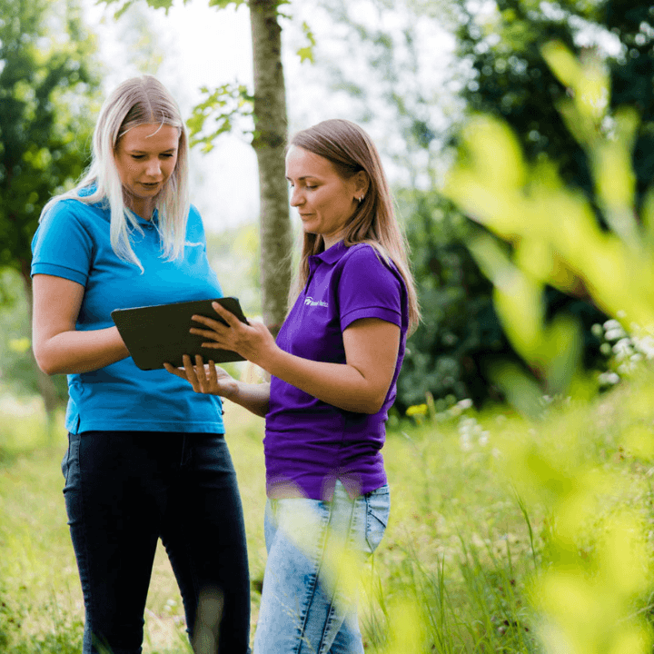 Twee glimlachende vrouwelijke zorgmedewerkers staan in een natuurrijke omgeving met een tablet in hun handen.