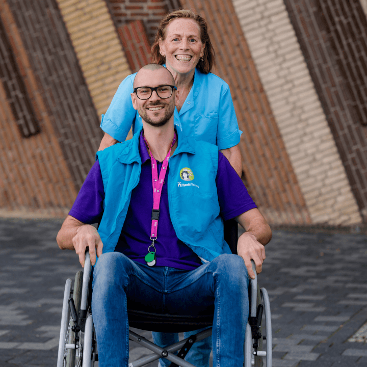 Twee lachende zorgverleners poseren buiten, waarbij een man in een rolstoel zit en een vrouw achter hem staat.