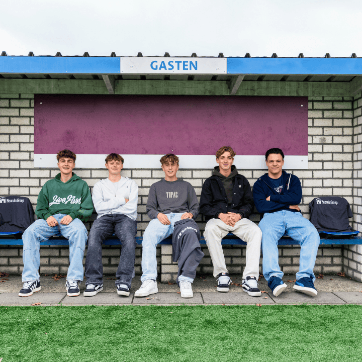Vijf jonge mannen zittend op een bankje onder een afdakje met bordje "GASTEN", grasveld op de voorgrond. Er liggen shirt van MeanderGroep op de stoelen naast hen.