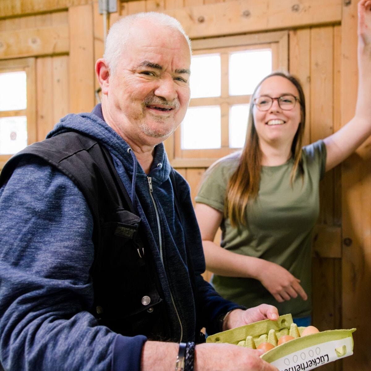 Een blije, oudere man houdt een doosje verse eieren vast. Een jonge vrouw staat glimlachend achter hem.