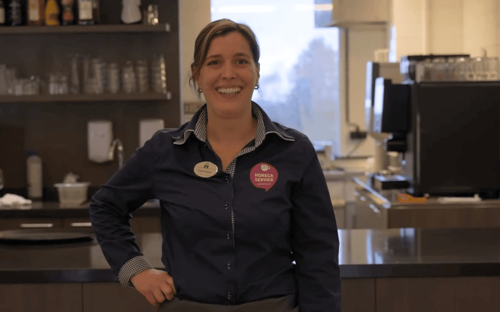 Vrouwelijk horecamedewerker met donkerblauw shirt en naamplaatje, glimlachend achter een bar of balie in een restaurant of café.