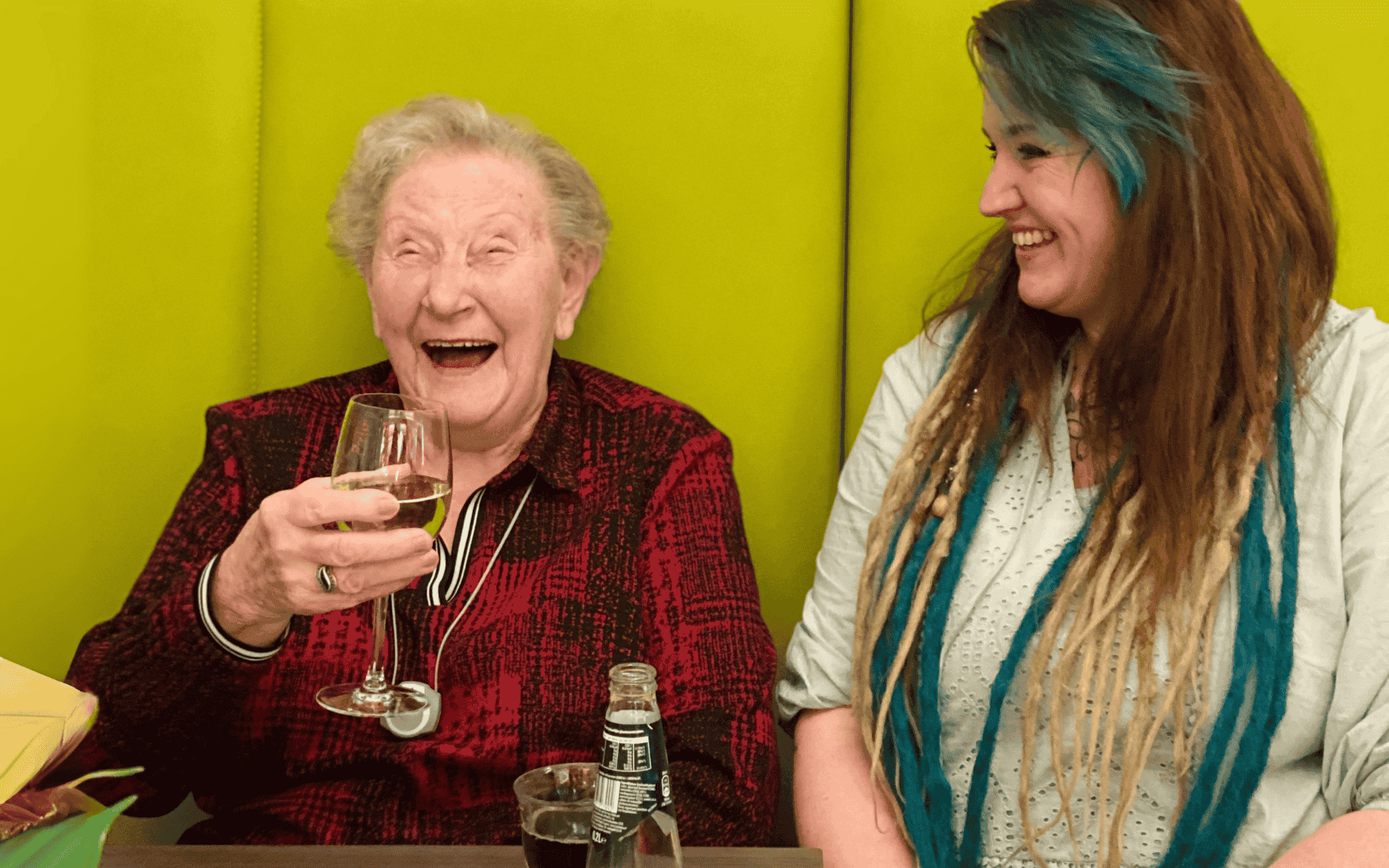Een oude lachende vrouw met een glas wijn zit naast een jonge lachende vrouw in een wonen met zorg woning.