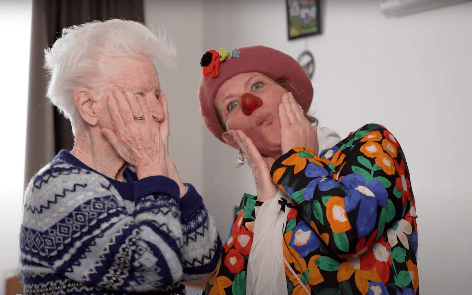Oude vrouw en clown met rode neus houden handen tegen hun gezichten en kijken speels naar elkaar.