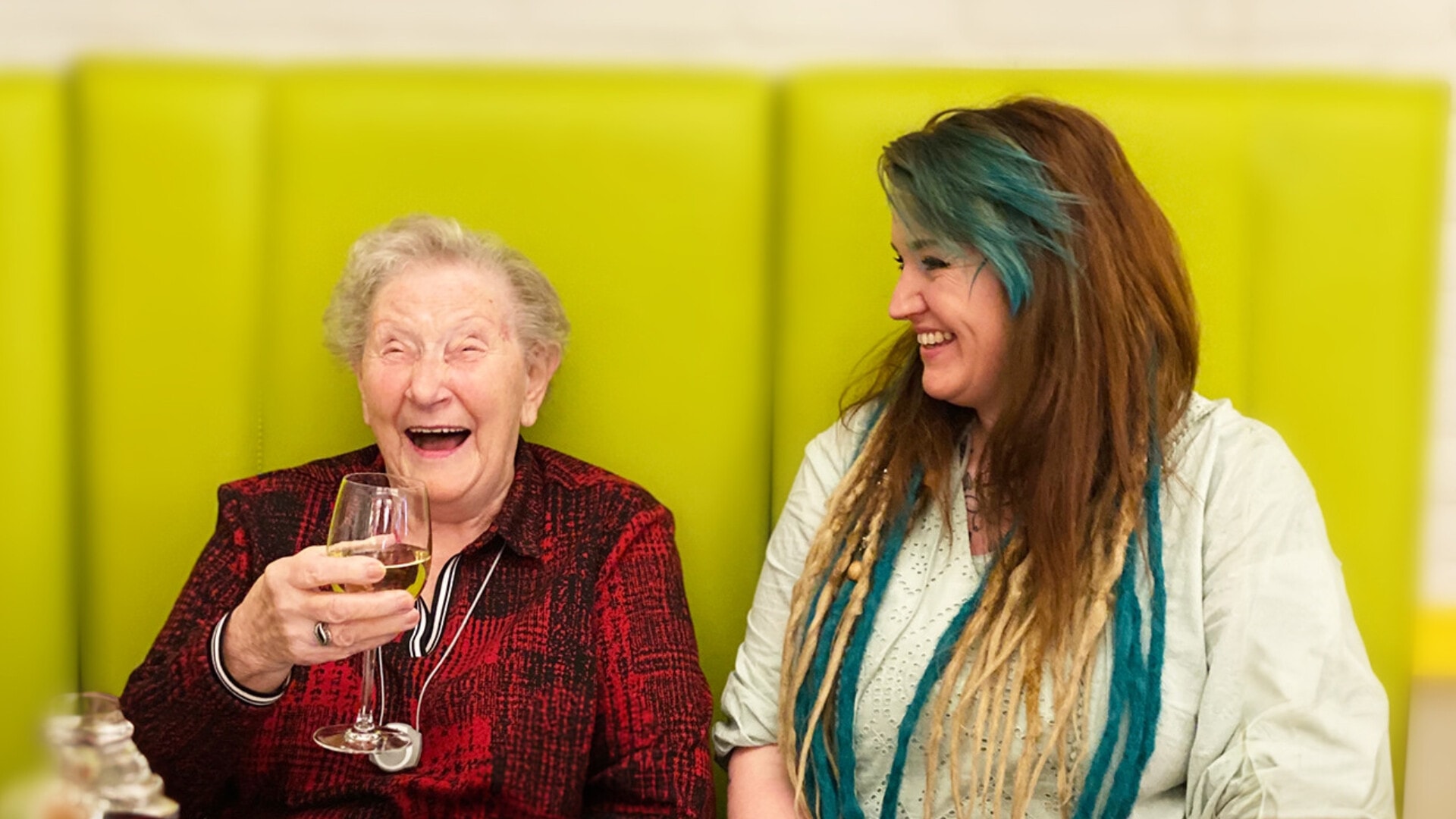 Oude lachende vrouw met rood bloesje en een glas wijn vast zit naast een jonge vrouw.