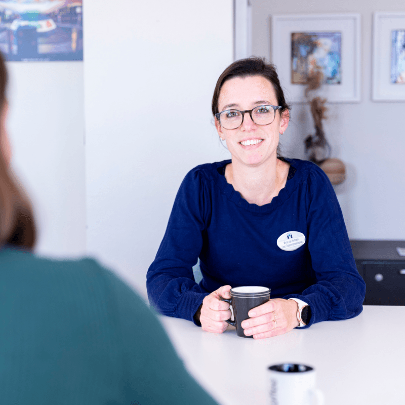Lachende vrouw met donkerblauwe trui kijkt recht in de camera en houdt een mok vast.