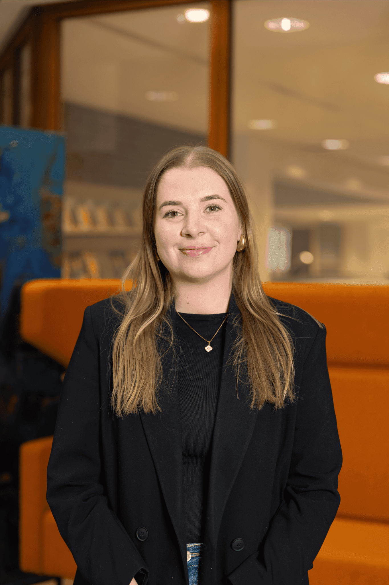 Jonge, vrolijke vrouw met een zwarte blazer staand voor een oranje bank in een modern kantoorinterieur.