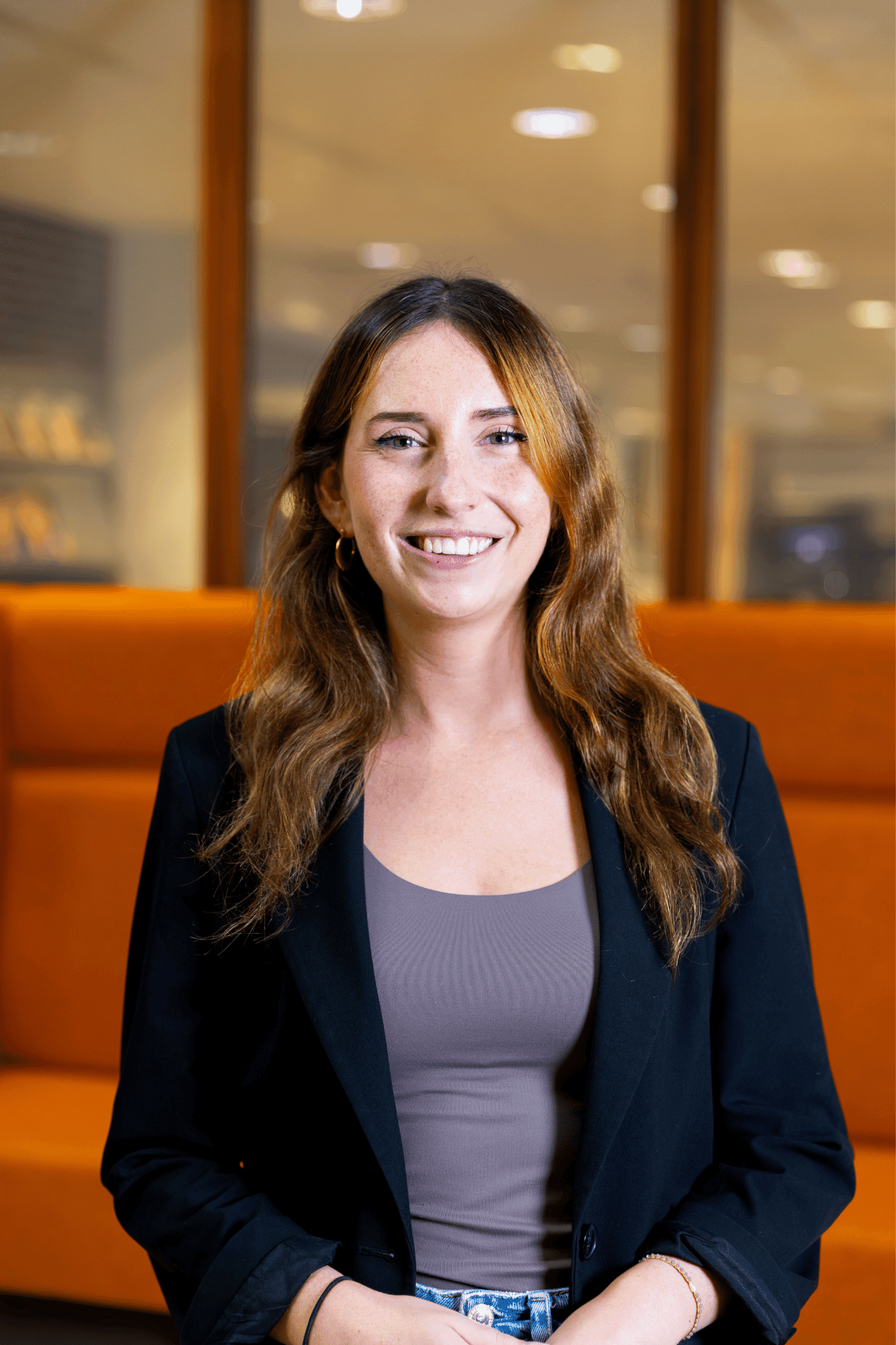 Jonge vrouw met lang bruin haar, lachend, gekleed in een zwart jasje en grijs shirt, zittend voor een oranje bank in een modern kantoorinterieur.