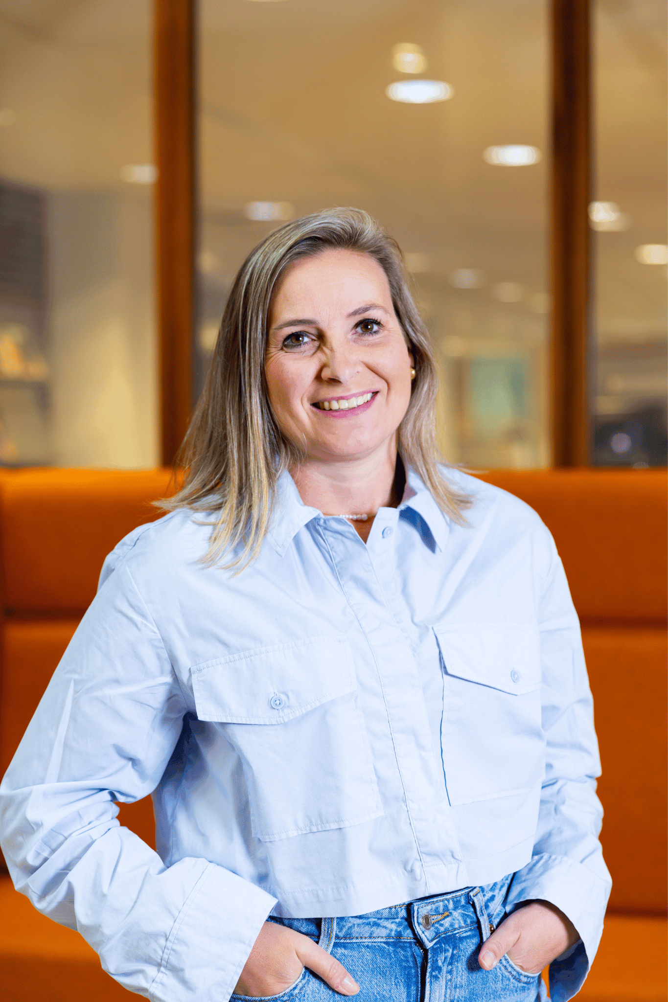 Poserende, lachende vrouw met lichtblauwe blouse staand voor een oranje bank in een modern kantoorinterieur.