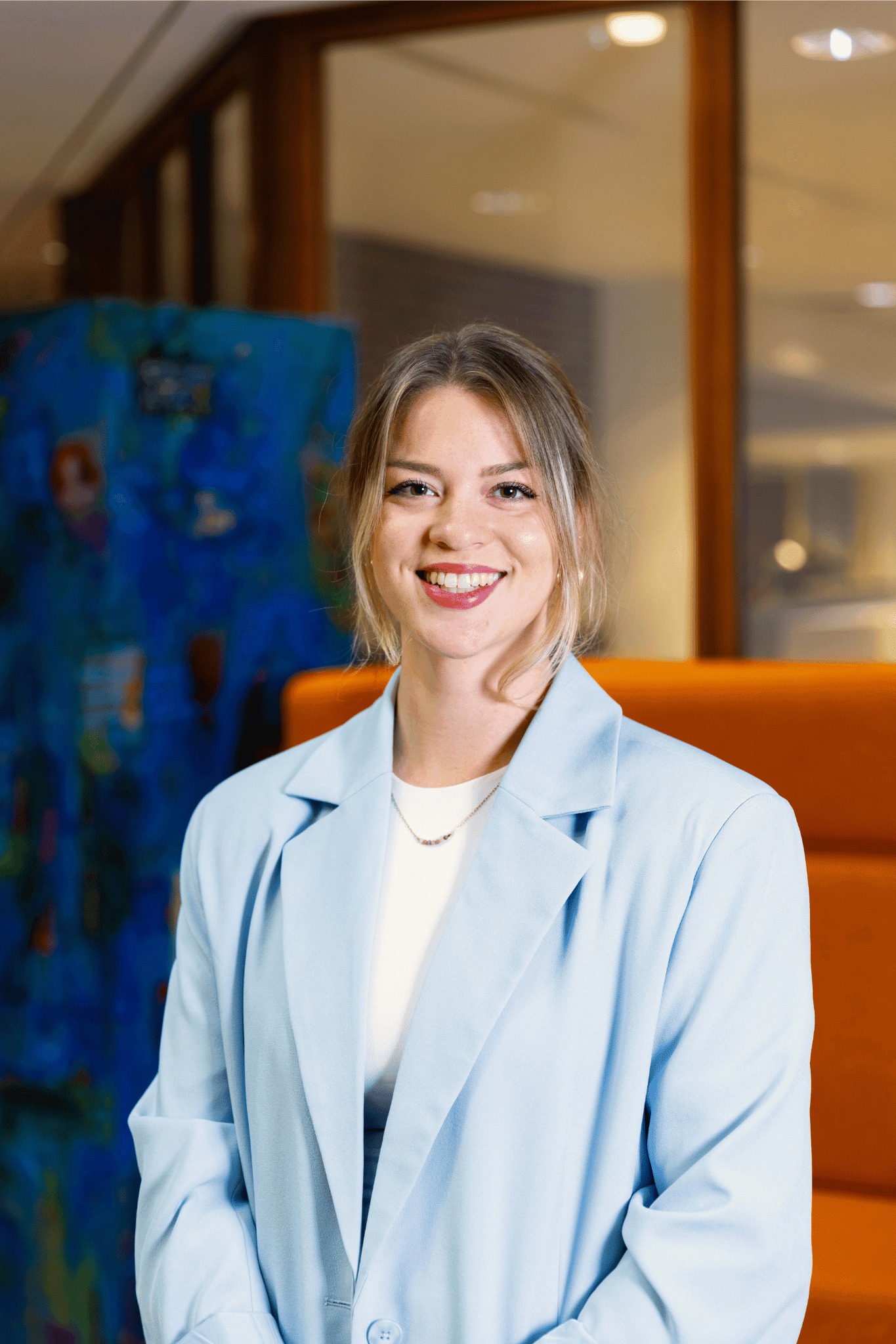 Jonge lachende vrouw met een lichtblauwe blazer staand voor een oranje bank in een modern kantoorinterieur.