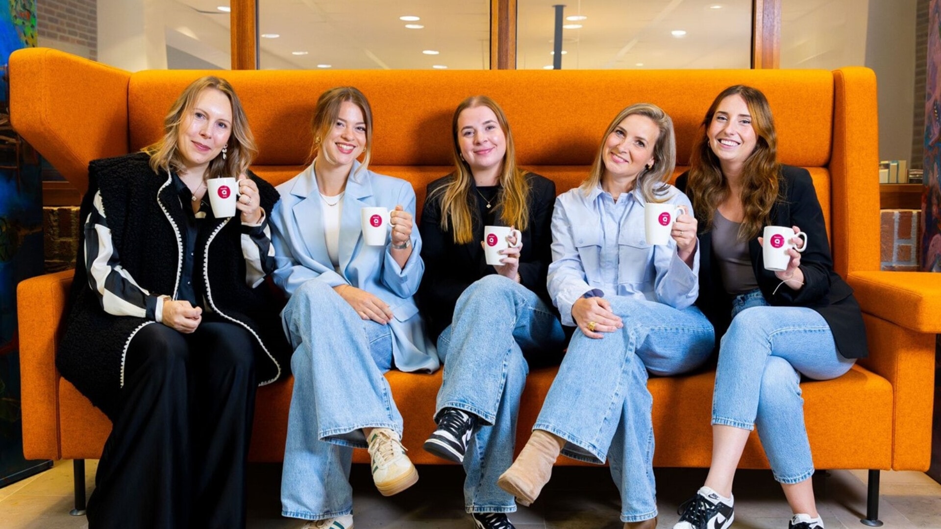 Vijf lachende vrouwen van Team Recruitement van MeanderGroep die op een feloranje bank zitten. De vrouwen houden witte mokken vast met een rood logo.