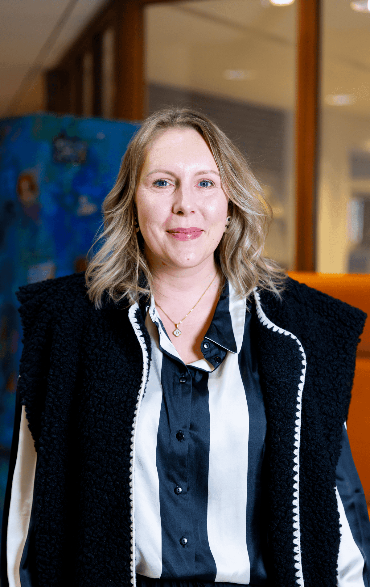 Portret van een vriendelijk lachende vrouw met halflang blond haar, gekleed in een zwart-wit gestreepte blouse en een donker gilet.